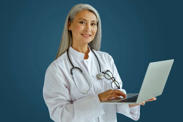 Χαμογελώντας ώριμη γυναίκα γιατρός σε λευκό παλτό λειτουργεί σε φορητό υπολογιστή στέκεται σε μπλε φόντο — Φωτογραφία Αρχείου