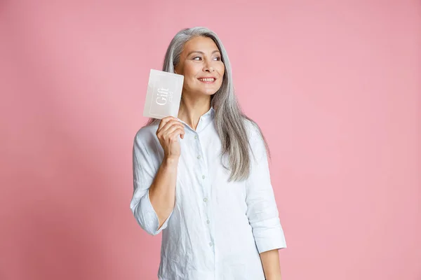 Pretty silver haired Asian woman holds gift card having ideas in studio — Stock Photo, Image