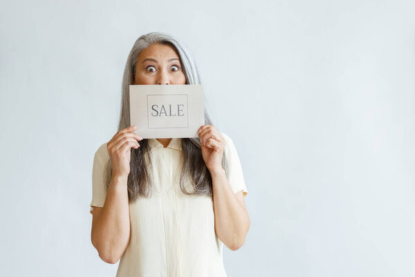 Shocked silver haired Asian lady holds card with word Sale on light grey background