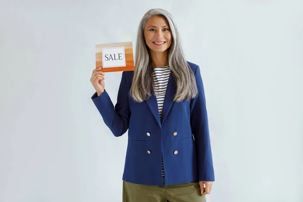 Hübsche asiatische Dame mit grauen Haaren hält Karte mit Wort Verkauf auf hellem Hintergrund — Stockfoto