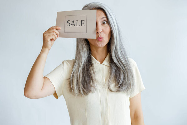 Funny mature woman with hoary hair covers eye with Sale sign on light grey background