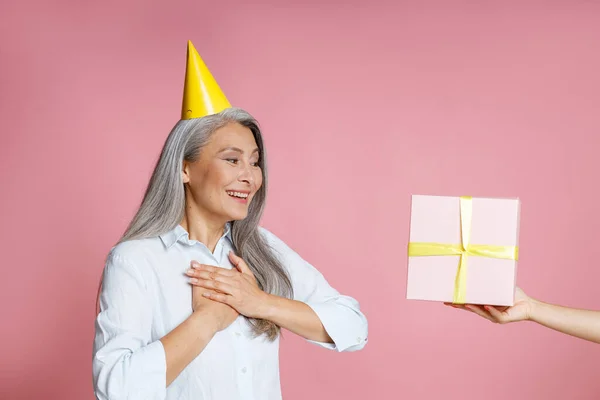 Medelålders asiatisk dam med fest hatt tittar på presentförpackning på vän hand i studio — Stockfoto