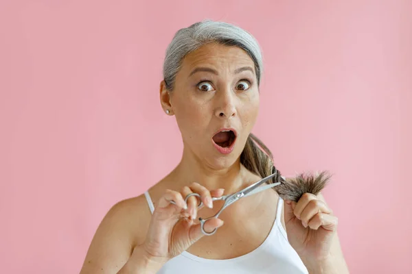 Geschokt middelbare leeftijd aziatische dame snijdt gespleten uiteinden van grijs haar op roze achtergrond — Stockfoto