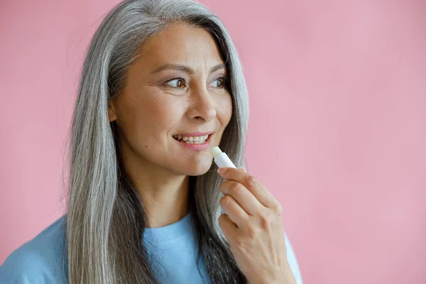 Positiva mujer asiática de pelo plateado aplica bálsamo labial sobre fondo rosa —  Fotos de Stock