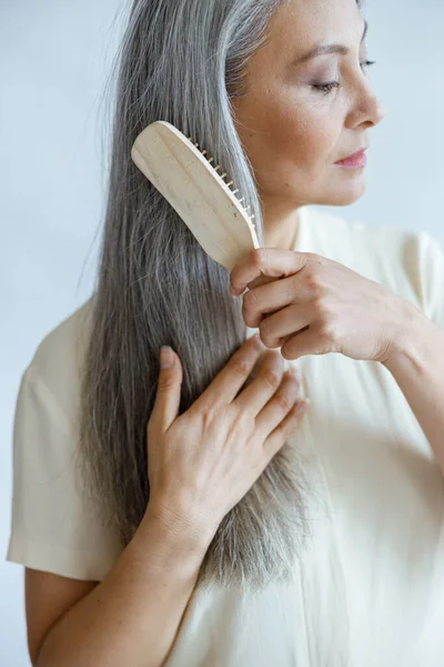 Tranquil middelbare leeftijd Aziatische model borstels lange taille haar poseren op lichte achtergrond — Stockfoto