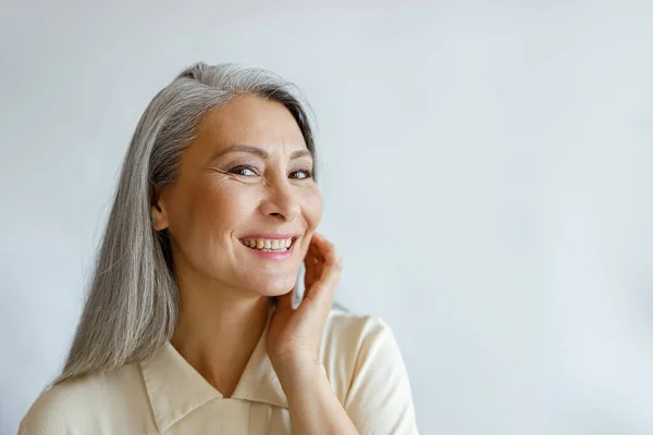 Atractiva mujer de pelo plateado en camisa beige toca mejilla sobre fondo gris claro —  Fotos de Stock