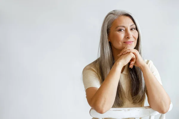 Happy middle aged woman with silver hair sits leaning on hands on light background — Stock Photo, Image