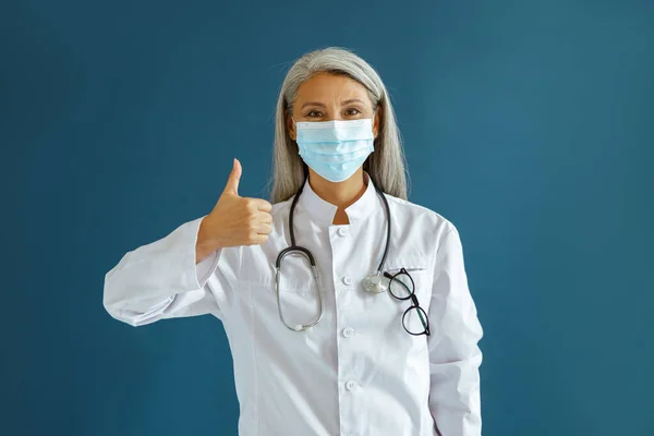 Θετική ώριμη Ασιάτισσα γιατρός με μάσκα δείχνει τον αντίχειρα επάνω στο μπλε φόντο — Φωτογραφία Αρχείου