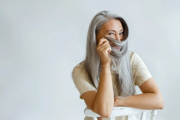 Positive middle aged Asian lady uses hoary hair as niqab sitting on chair in studio — Stock Photo, Image