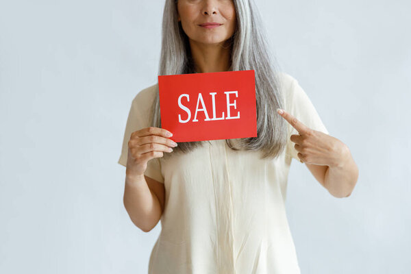 Lady with long grey hair holds red Sale sign pointing by finger on light background