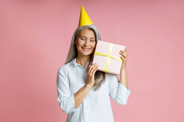Happy mature Asian lady with yellow party hat holds gift box on pink background — Stock Photo, Image