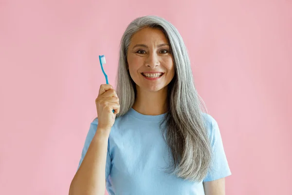 Alegre meia idade ásia mulher detém escova de dentes de pé no rosa fundo — Fotografia de Stock