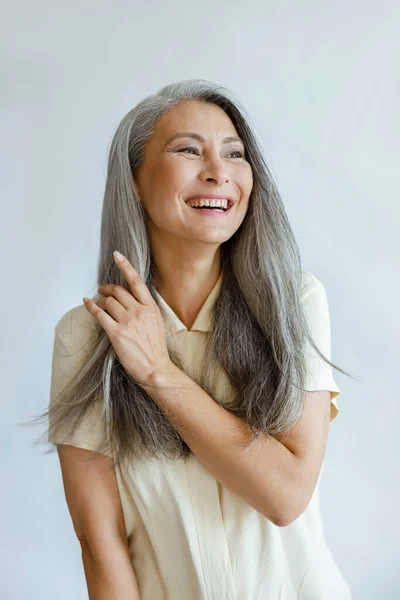 Middle aged Asian woman touches natural hoary hair on light grey background — Stock Photo, Image