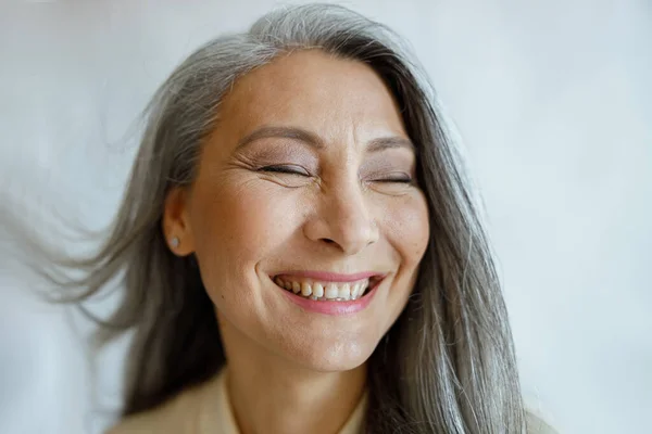 Joyeuse modèle féminin asiatique d'âge moyen avec des cheveux argentés volants sur fond gris clair — Photo