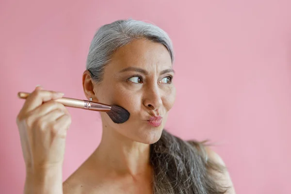 Komik beyaz saçlı Asyalı kadın pembe arka planda fırçayla makyaj yapıyor. — Stok fotoğraf