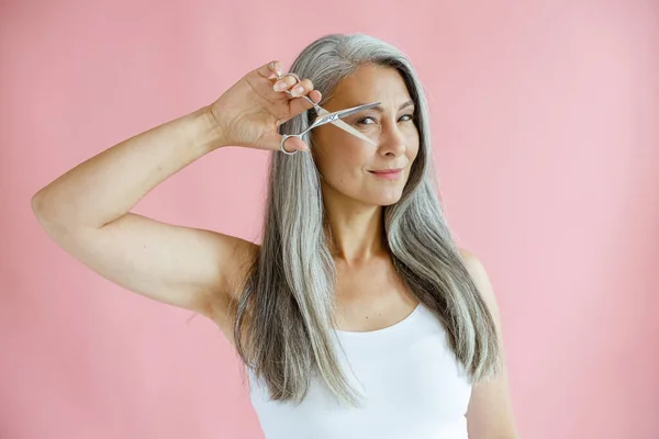 Beautiful Asian lady with grey hair holds open scissors near face on pink background — Stock Photo, Image