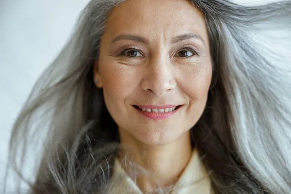 Middle aged Asian woman with flying hoary hair looks into camera on light grey background — Stock Photo, Image