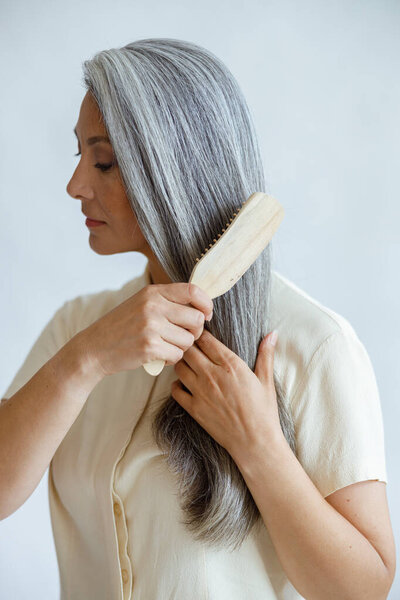 Languid middle aged Asian woman brushes natural hoary hair on light grey background — Stock Photo, Image