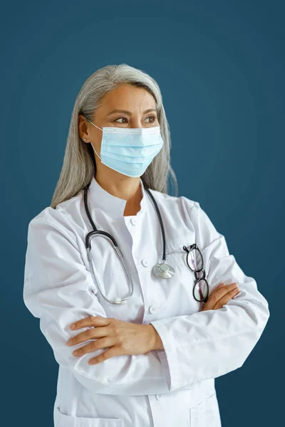 Hoary μαλλιά Ασιάτισσα γιατρός γυναίκα με μάσκα και σταυρωμένα χέρια στέκεται σε μπλε φόντο — Φωτογραφία Αρχείου