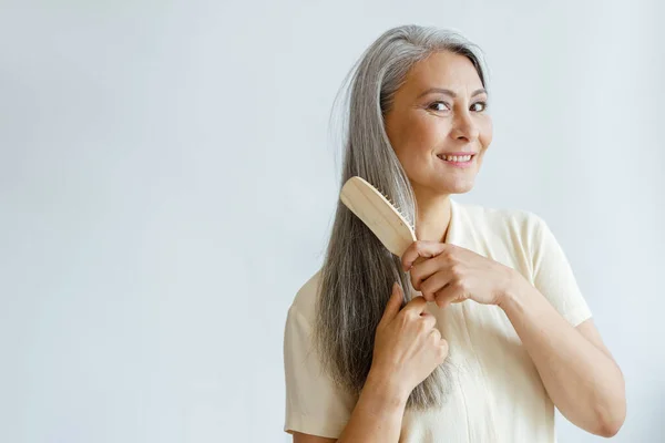 Bedachtzame middelbare leeftijd aziatische vrouw borstels grijs haar poseren op lichte achtergrond — Stockfoto