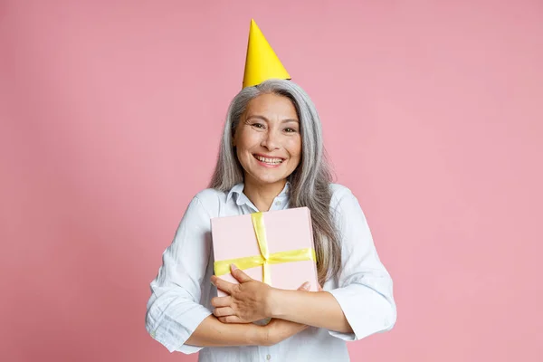 快乐的亚洲女人，头发灰白，头戴亮晶晶的派对帽，在工作室里拿着礼品盒 — 图库照片