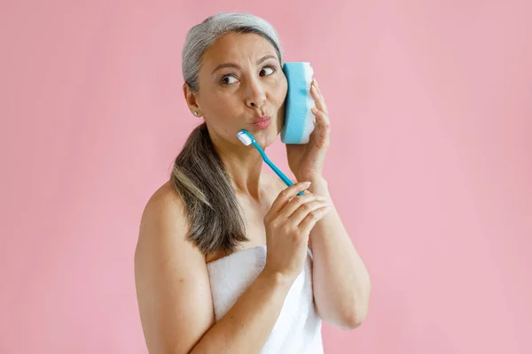 Mediana edad señora asiática con toalla de baño sostiene cepillo de dientes usando esponja como teléfono en el estudio —  Fotos de Stock