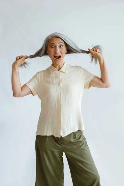 Shocked Asian woman holds hoary hair locks on light grey background — Stock Photo, Image