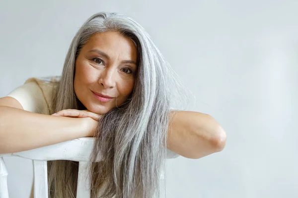 Cheerful middle aged Asian lady with flowing hoary hair sits backwards on chair — Stock Photo, Image