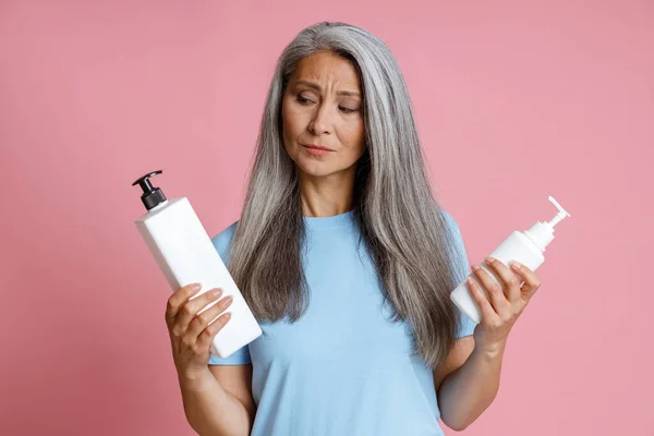 Nachdenkliche Asiatin mittleren Alters wählt Kosmetikprodukt auf rosa Hintergrund — Stockfoto