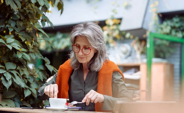 Grey haired senior woman eats delicious dessert sitting at table outdoors — Stock Photo, Image