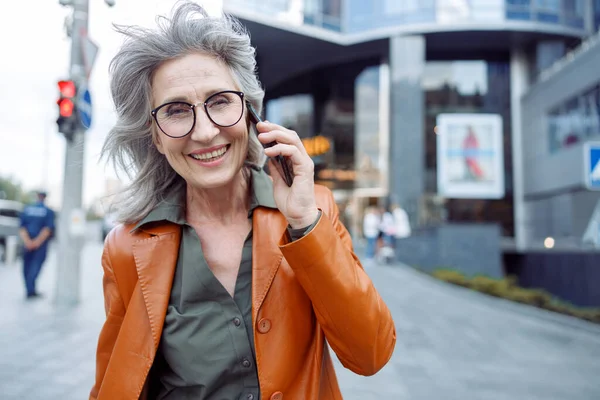 Happy grey haired senior woman with glasses talks on mobile phone on city street — Stock Photo, Image