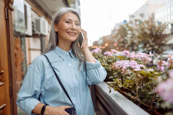 Positive mature Asian lady in blue shirt talks on mobile phone on terrace with flowers — Stock Photo, Image