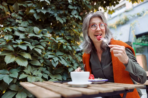 Cheerful senior woman with spectacles eats strawberry dessert sitting at table on outdoors cafe terrace — Stock Photo, Image