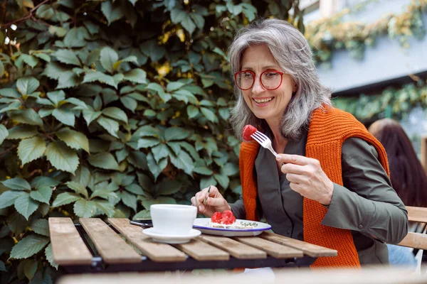 Smiling mature female guest with glasses eats toast at table on outdoors cafe terrace — Stock Photo, Image