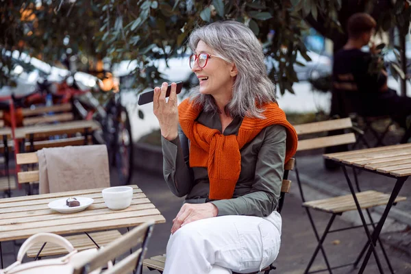 Silver haired lady with glasses uses loudspeaker mode on phone on outdoors cafe terrace — Stock Photo, Image