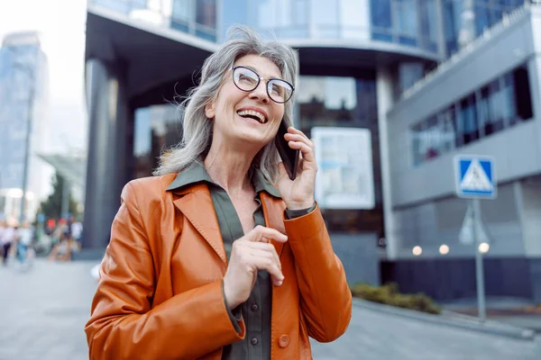 Joyful hoary haired senior lady talks on cellphone on modern city street — Stock Photo, Image
