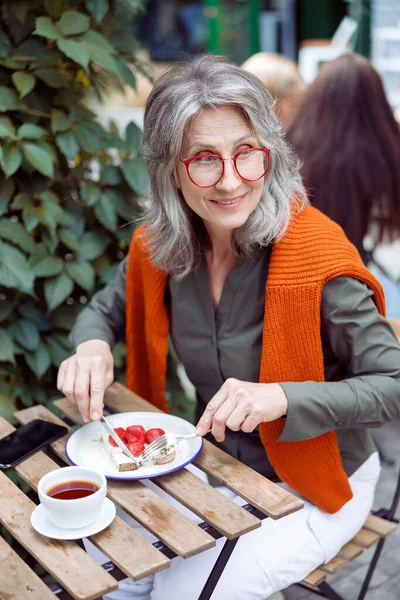Smiling mature lady eats toast with cream and strawberries sitting at table on outdoors cafe terrace — Stock Photo, Image