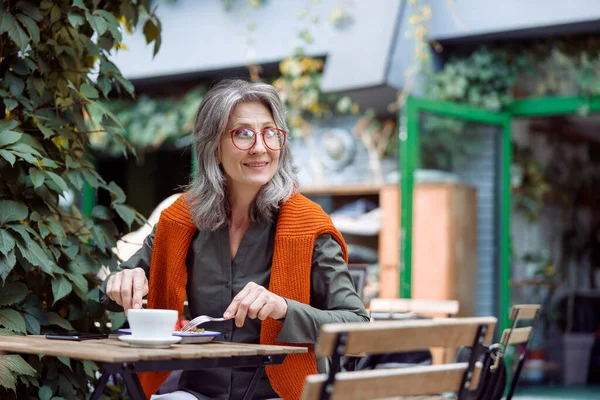 Happy mature woman with glasses eats dessert sitting at small table outdoors — Stock Photo, Image