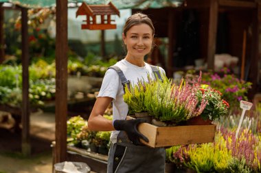 Neşeli bir kadın neşeyle bir bahçe merkezinde içi canlı çiçeklerle dolu ahşap bir sandık tutuyor.