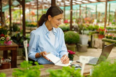 Bir kadın görevlerini etkin bir şekilde canlı, renkli bir sera ortamında dizüstü bilgisayarını ve defterini kullanarak düzenler.
