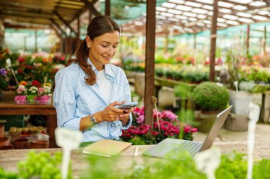 Renkli bir çiçekçide çalışan bir kadın dizüstü bilgisayarı ve akıllı telefonuyla verimliliğini ve yaratıcılığını arttırıyor.