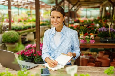 Neşeli bir kadın, çeşitli bitkilerle dolu canlı bir çiçekçinin içine mutlu bir şekilde renkli notlar yazar.