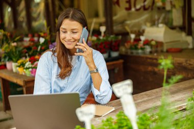 Mutlu, gülümseyen bir kadın, canlı bir çiçekçide bilgisayarını kullanırken bir telefon görüşmesinde ustaca birden fazla iş yapar.