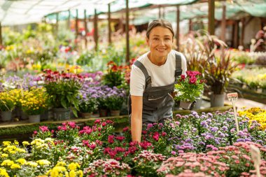 Neşeli bir kadın bir bahçe merkezinde renkli çiçeklerin arasında gururla poz verir, doğaya olan tutkusunu gösterir.