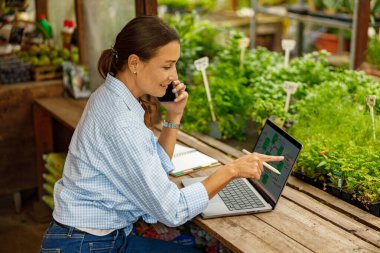 Bir kadın akıllı bir şekilde bilgisayarını ve telefonunu canlı bir serada kullanır, doğayı teknolojiyle harmanlar.