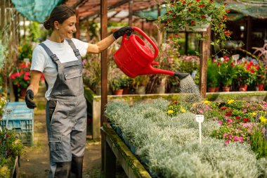 Renkli tulumlu bir kadın çiçeklerle çevrili parlak bir serada çiçekleri mutlu bir şekilde suluyor.