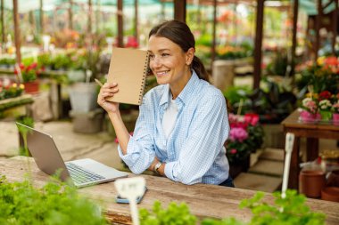 Canlı bir çiçekçide çalışırken, dizüstü bilgisayarını ve not defterini kullanarak mutlu bir şekilde gülümseyen bir kadın.