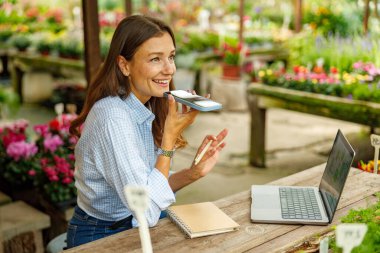 Neşeli bir kadın, canlı bir bahçe dükkanında çiçek açan çiçeklerle çevriliyken mutlu bir sese bağlanır.