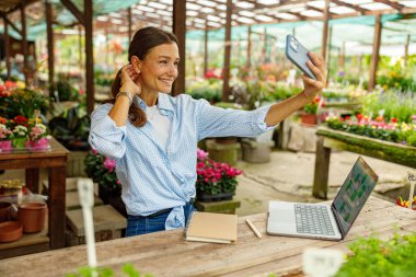 Neşeli ve neşeli bir kadın, güzel çiçeklerle çevrili güzel bir bahçe merkezinde canlı bir selfie çeker.