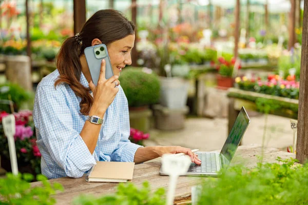 Bir kadın dizüstü bilgisayarında çalışıyor ve canlı çiçeklerle dolu renkli bir serada telefonla konuşuyor.